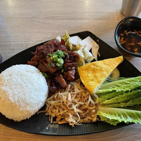 Vietnamese grilled pork with rice, fried onions, lettuce, and a side of soup on a black plate.