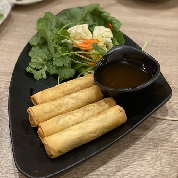 Four golden-brown Vietnamese spring rolls with a side of fresh greens and dipping sauce on a black plate.
